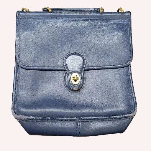 A blue leather handbag with a top handle and a front flap secured by a snap button, resting on a piece of brown paper on a wooden surface.