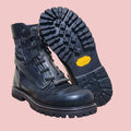 A pair of black leather lace-up boots displayed on a wooden stool, with one boot standing upright and the other showing its treaded sole with a yellow Vibram logo.