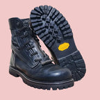 A pair of black leather lace-up boots displayed on a wooden stool, with one boot standing upright and the other showing its treaded sole with a yellow Vibram logo.