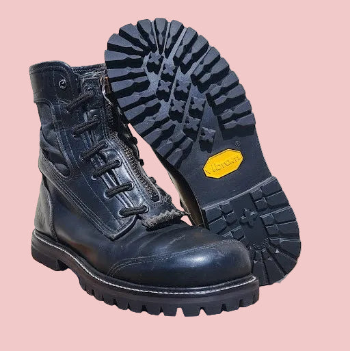 A pair of black leather lace-up boots displayed on a wooden stool, with one boot standing upright and the other showing its treaded sole with a yellow Vibram logo.