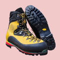 A pair of yellow and black high-top climbing boots displayed on a wooden stool, with one boot standing upright and the other showing its textured black outsole.