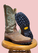 A pair of western boots displayed on a wooden stool against a textured wall. One boot stands upright showing its tan leather shaft with light blue embroidery and brown foot, while the other lies on its side displaying a black treaded sole with a small yellow logo.