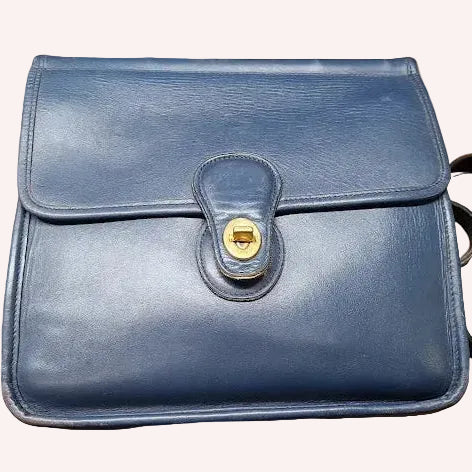 A blue leather shoulder bag with a flap closure and a gold-tone snap button, shown against a solid pink background.