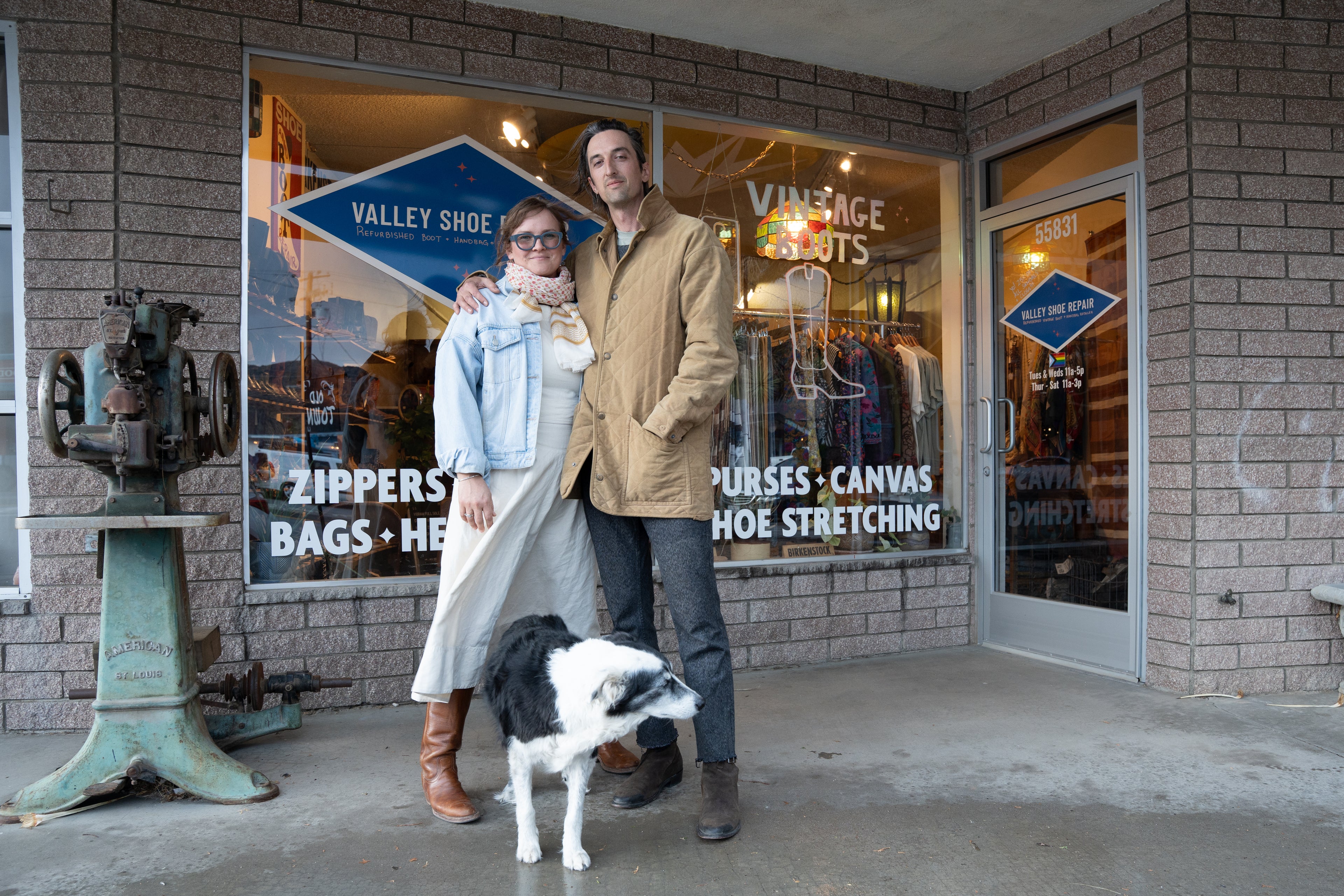 Caileen and Birdie stand in front of their shoe repair shop with a black and white dog at their feet