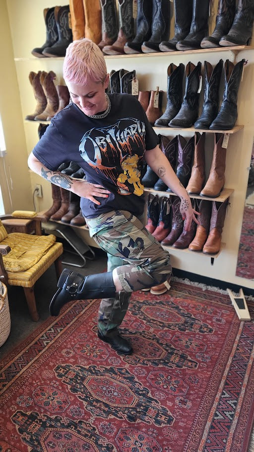A person with short pink hair poses in a boot shop, wearing a black graphic t-shirt, camouflage cargo pants, and black boots. Shelves behind them are filled with various cowboy boots, and a red patterned rug covers the floor.