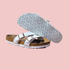 A pair of Birkenstock Arizona slide sandals with white straps and brown footbeds, displayed on a wooden stool against a textured light gray wall.