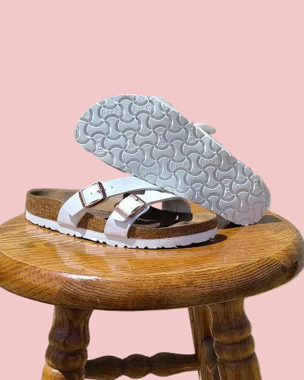 A pair of Birkenstock Arizona slide sandals with white straps and brown footbeds, displayed on a wooden stool against a textured light gray wall.