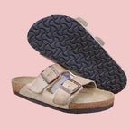 A pair of beige double-strap slide sandals with brown buckles and cork soles, displayed on a wooden stool with one sandal showing its black treaded sole against a light beige wall.