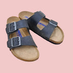 A pair of dark gray double-strap leather sandals with silver buckles and tan footbeds, displayed on a round wooden stool.