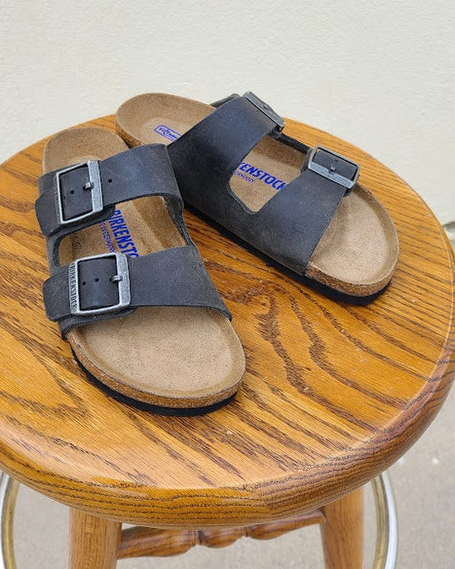 A pair of dark gray double-strap leather sandals with silver buckles and tan footbeds, displayed on a round wooden stool.