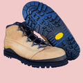 Tan work boots with blue soles on a wooden stool against a white brick wall.