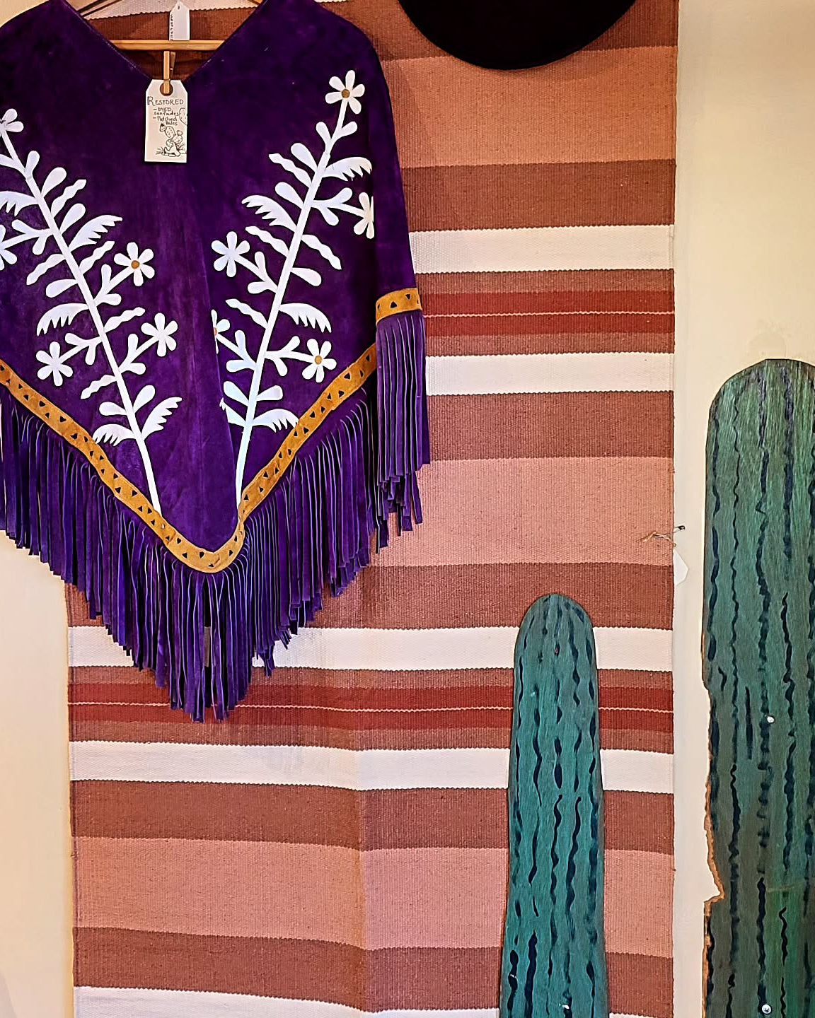 Purple poncho with white patterns and gold fringe on a striped wall with cacti.